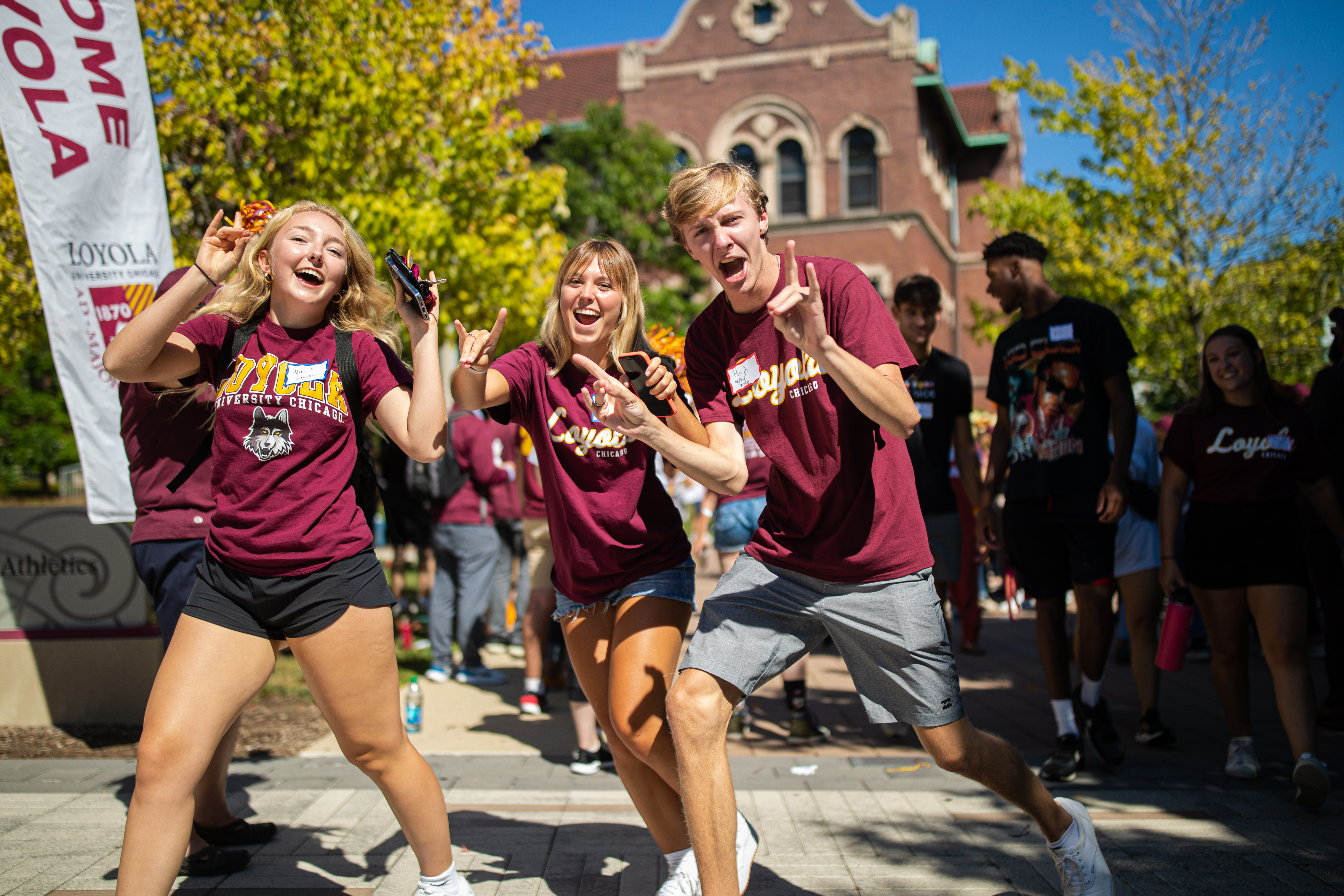 Three students in a crowd wearing Loyola University Chicago t-shirts at Freshmen Orientation, laughing and giving the peace sign, as they walk through the Lake Shore campus.
